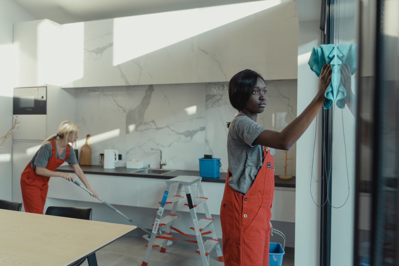 journey Two women cleaning a modern kitchen, wiping glass surfaces and mopping the floor.